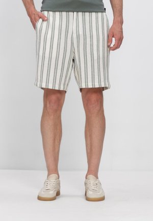 Man met witte korte broek met verticale zwarte strepen en witte sneakers staand tegen een effen achtergrond.