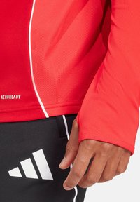 Maglietta atletica rossa a manica lunga con texture a rete, dettagli in piping bianco e logo sul bordo inferiore. Pantaloni neri con un logo bianco ben visibile.