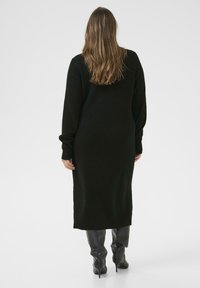Langes schwarzes Strickkleid mit lässiger Passform, abgesenkten Schultern und gerippten Bündchen, kombiniert mit schwarzen kniehohen Stiefeln mit Absatz.