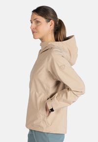 Beige vattentät jacka med huva, som har en framdragkedja och fickor. Tillverkad av mjukt, lätt material med en figursydd design.