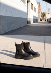 Paire de bottines en cuir noir à semelles épaisses posées sur un trottoir urbain ensoleillé près d’un bâtiment en briques gris et blanc.