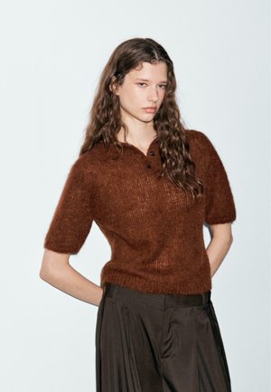 Brauner, kurzärmeliger Strickpullover aus weichem, strukturiertem Material, mit Kragen und zwei schwarzen Knöpfen, kombiniert mit dunkel gestreiften Hosen.