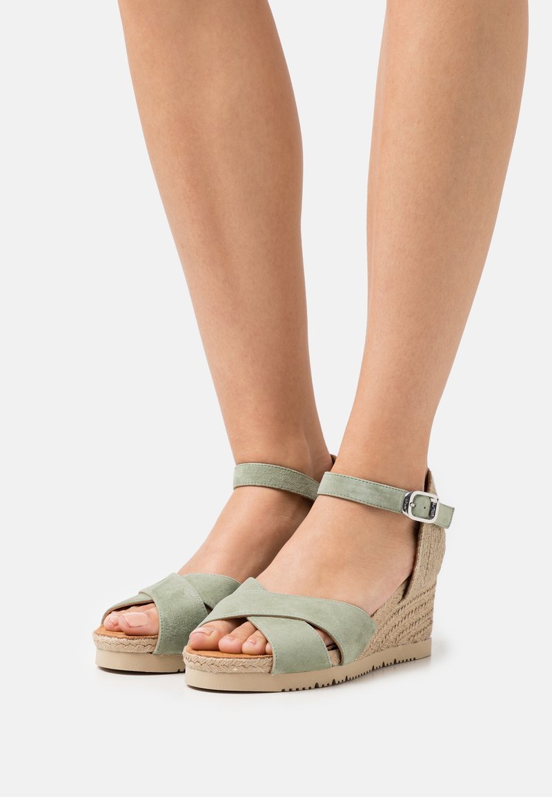 Sandales compensées en suède vert clair avec brides à la cheville et détails tressés portées pieds nus sur un fond uni.