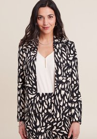 Blazer a righe bianche e nere con un design aderente, abbinato a una maglietta bianca con una zip discreta. Tessuto texturizzato con cuciture visibili.