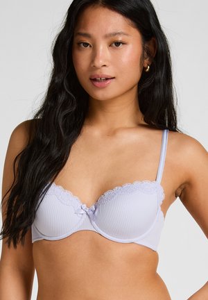 Jeune femme aux longs cheveux noirs portant un soutien-gorge violet clair orné de dentelle, posant devant un fond gris clair uni.