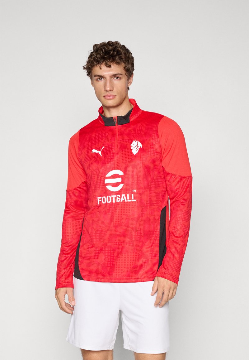 Mann iført rød langermet sportstrøye med logoer og hvite shorts, stående mot en enkel lys bakgrunn.