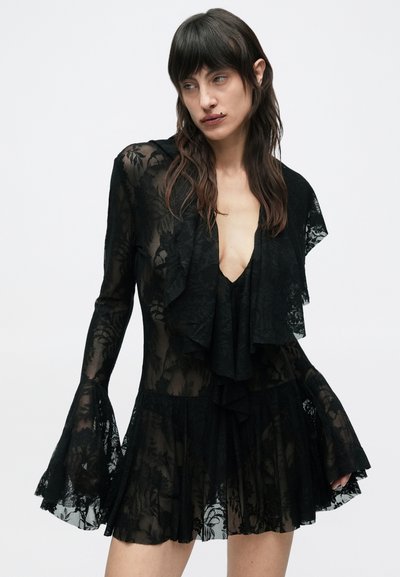 Γυναίκα που φοράει μαύρο διάφανο δαντελένιο φόρεμα με μακριά μανίκια καμπάνα και βαθιά V λαιμόκοψη, στέκεται μπροστά σε απλό λευκό φόντο.