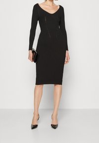Abito nero a coste con maniche lunghe, scollatura a V, vestibilità aderente, lunghezza al ginocchio, delicato taglio in vita, abbinato a una pochette nera.