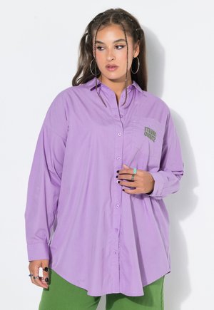 Young woman with braided hair wearing hoop earrings, oversized lilac button-up shirt, and green pants, looking to the side against a white background.
