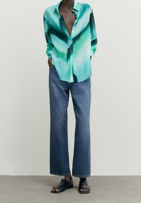 Chemise boutonnée en dégradé de bleu sarcelle, associée à un jean large en denim bleu. Sandales noires complètent la tenue. Tissu lisse avec une coupe décontractée.