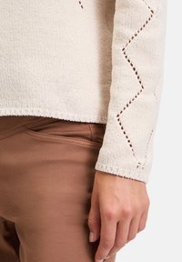 Ljus beige texturerad tröja med zickzackutskärning på ärmarna, tillsammans med en brun slimmad byxa, som framhäver handpositionen.