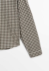 Långärmad skjorta med brunt och vitt rutmönster i gingham, visar manschetten och en del av fållen mot en vit bakgrund.