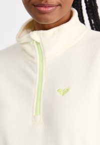 Quarter-zip fleece pullover in cream with a textured surface. Features a light green zipper and heart-shaped logo in matching green embroidery.