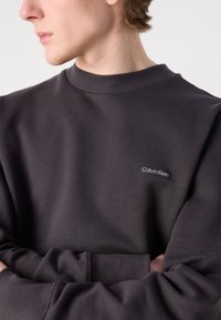 Temno siva majica s kapuco z rebrastim ovratnikom in manšeti. Ima majhen črn etiket s napisom "Calvin Klein" spredaj. Mehka, teksturirana tkanina.