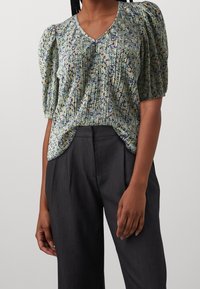Blusa de manga corta con estampado floral y escote en V. Textura plisada, con tonos de verde, púrpura y amarillo. Combinada con pantalones oscuros.