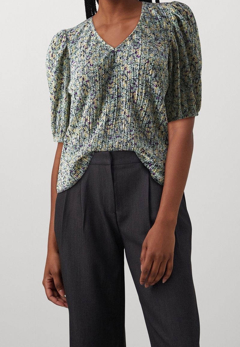 Blusa de manga corta con estampado floral y escote en V. Textura plisada, con tonos de verde, púrpura y amarillo. Combinada con pantalones oscuros.