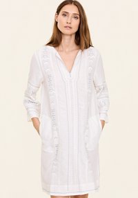 Weißes Tunika-Kleid mit langen Ärmeln, V-Ausschnitt, Fronttaschen und aufwendiger hellblauer Stickerei entlang der Vorderseite und Ärmeln.
