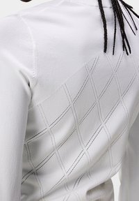 Plano cercado de una camisa blanca de manga larga con un sutil patrón de rombos bordado en la parte posterior, usada por una persona con cabello trenzado.