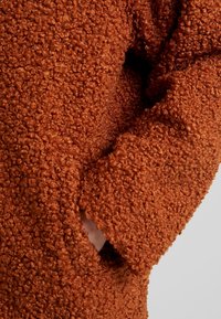 Texturerad orange stickad tröja med krullig fleece. Har en lös passform och en sidficka, vilket betonar värme och komfort.