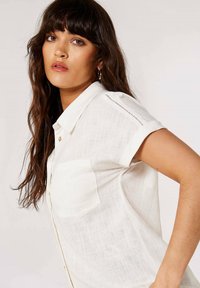 White linen shirt with short rolled sleeves, front pocket, and decorative stitching on the shoulder. Lightweight and airy texture.