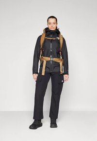 Veste noire imperméable avec capuche et fermeture éclair, associée à un pantalon cargo noir et des chaussures de randonnée noires, accompagnée d'un sac à dos marron.