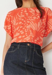 Blusa color coral con un patrón floral, mangas cortas amplias y un corte relajado, combinada con una falda de mezclilla oscura con costuras naranjas.