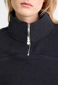 Schwarzer Fleece-Pullover mit hohem Kragen, ausgestattet mit einem halben Reißverschluss und einem Perlenakzent am Reißverschluss-Zipper. Weiche Textur und schlichtes Design.