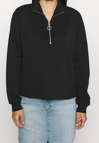 Svart zip-up sweatshirt med ribbad krage och frampanel, med en silverdragkedja. Modellen matchar den med en ljusblå jeanskjol.