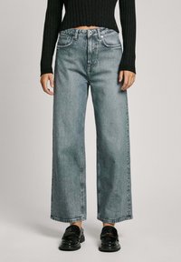 Ljusblå vida jeans i denim, med framfickor, knäppning och synliga sömmar. Svarta skor med glänsande yta.