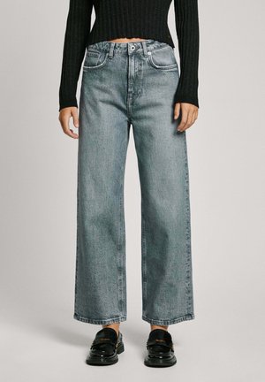 Hellblaue, weit geschnittene Jeans aus Denim mit vorderen Taschen, Knopfverschluss und sichtbaren Nähten. Schwarze Schuhe mit glänzender Oberfläche.