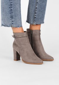 Botines de ante gris con punta afilada, cierre de cremallera y acentos de tachuelas en una correa. El tacón es de forma bloque, proporcionando estabilidad.