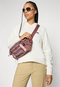 Ungdom som har solbriller på, hvit hettegenser, beige bukser, holder en mauve Patagonia crossover-veske mot en ensfarget bakgrunn.