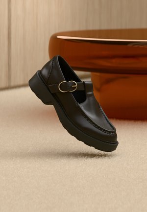 Mocassino in pelle nera con cinturino con fibbia dorata, suola spessa, mostrato fluttuante sopra un tappeto beige con un tavolo di legno marrone sullo sfondo.