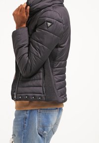 Svart vadderad jacka med hög krage, ribbade sidopaneler, dragkedjefickor och tryckknappar vid fållen. Har en logotyp på axeln.