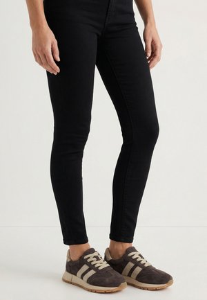 Persoon die zwarte slim-fit broek en bruine suède sneakers met beige veters en strepen draagt, staande op een witte vloer.