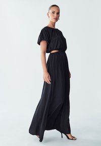 Gaun maxi hitam dengan lengan puff pendek dan potongan tengah di pinggang. Kain mengalir dengan tekstur halus, dipadukan dengan sandal bertumit tinggi.