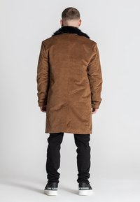 Abrigo largo de ante marrón con cuello de piel sintética negra, textura suave y corte recto, combinado con pantalones negros y zapatillas blancas.