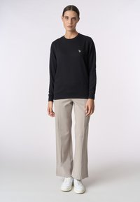 Felpa nera con maniche lunghe e polsini a coste, caratterizzata da un piccolo logo ricamato. Abbinata a pantaloni beige a gamba larga e sneakers bianche.
