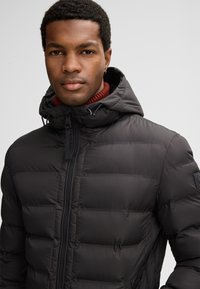 Giacca imbottita nera con cappuccio, dotata di chiusura con zip e tasche laterali. Superficie testurizzata con un design moderno e lineare.