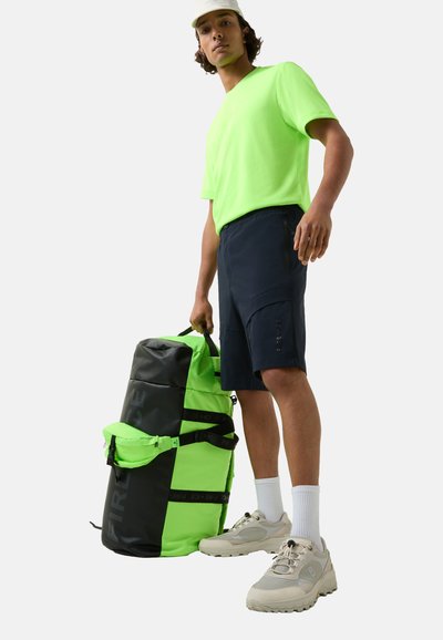 Neon grøn duffel taske med sorte detaljer og stropper, der har en struktureret overflade. Model iført lys T-shirt, mørke shorts og beige sko.
