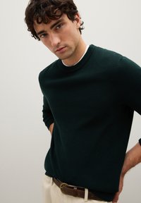 Maglione in un verde scuro e tessuto ruvido con collo tondo, indossato sopra una camicia bianca, abbinato a pantaloni chiari e una cintura marrone.