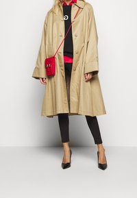 Khaki, oversize płaszcz trench z dużymi guzikami i szerokimi rękawami. Połączony z czarnymi legginsami i czarnymi szpilkami. Czerwona torebka na ramię.