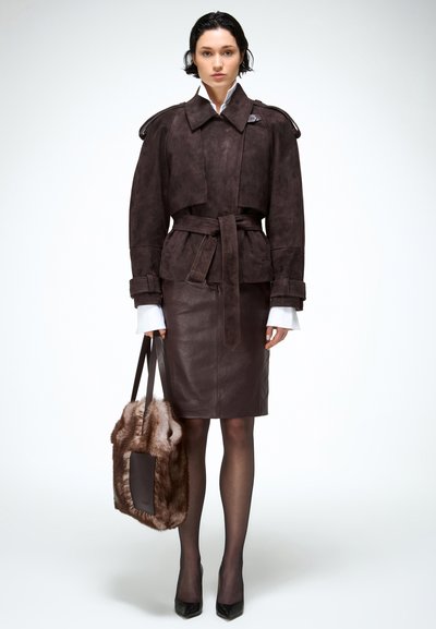 Nő sötétbarna öves dzsekiben és szoknyában, áttetsző fekete harisnyával, fekete magassarkú cipőben, barna szőrme díszítésű táskával a kezében, fehér háttér előtt áll.