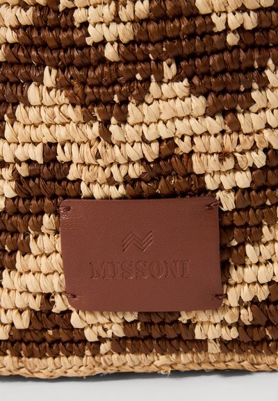 Κοντινό πλάνο από καφέ και μπεζ υφαντό υλικό με μια ραμμένη καφέ δερμάτινη ετικέτα που φέρει το ανάγλυφο "MISSONI" και ένα λογότυπο ζιγκ ζαγκ.