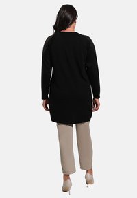 Black long-sleeve tunic with a textured knit pattern on the shoulders, paired with beige pants and beige heels, viewed from the back.