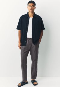 Short-sleeve navy button-up shirt over a white tank, paired with gray textured pants and black sandals. Simple design, relaxed fit.
