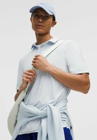 Jasnoniebieska koszulka polo o miękkiej fakturze, w zestawie z jasnoniebieską czapką. Przy boku leży torebka w kolorze kremowym, a w pasie związany jest jasny sweter.