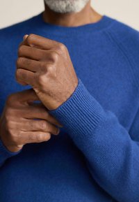 Blauer Strickpullover mit gerippten Bündchen, der eine glatte Textur und einen enganliegenden Schnitt aufweist. Hände, die den Ärmel nahe dem Handgelenk anpassen.