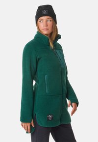 Giacca in pile verde con colletto alto, tasca sul petto con zip e fessure laterali. Cappellino nero con logo. Tessuto morbido e vestibilità rilassata.