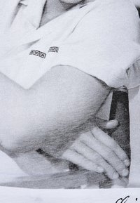 Zbliżenie białej tkaniny o subtelnej fakturze, z misternym czarnym geometrycznym wzorem na rękawie. Ręce są częściowo widoczne.